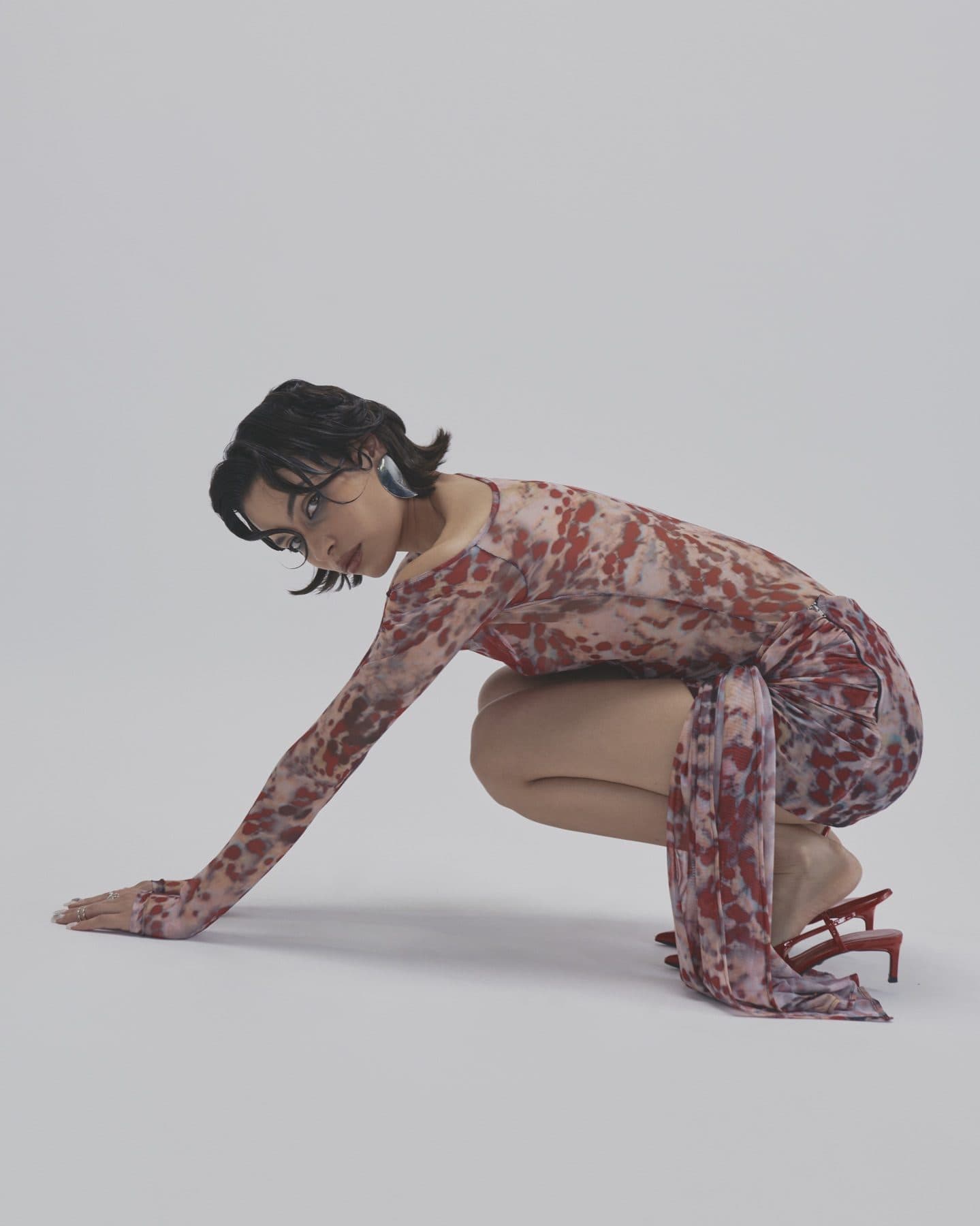 A model in a patterned long-sleeved dress crouches with one hand on the floor against a pale grey backdrop.