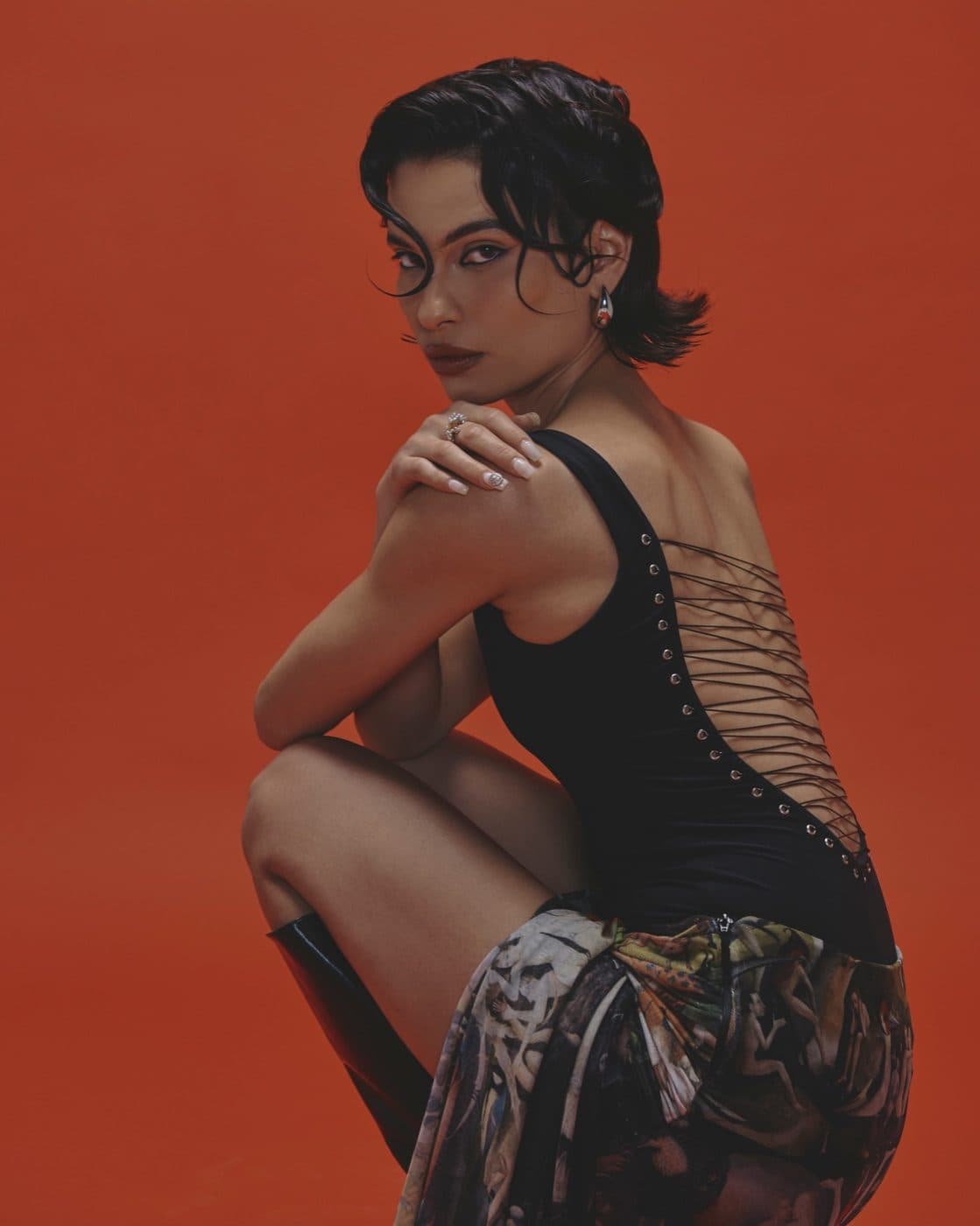A model in a black lace-up top and a patterned skirt crouches against a burnt orange backdrop, looking over her shoulder.