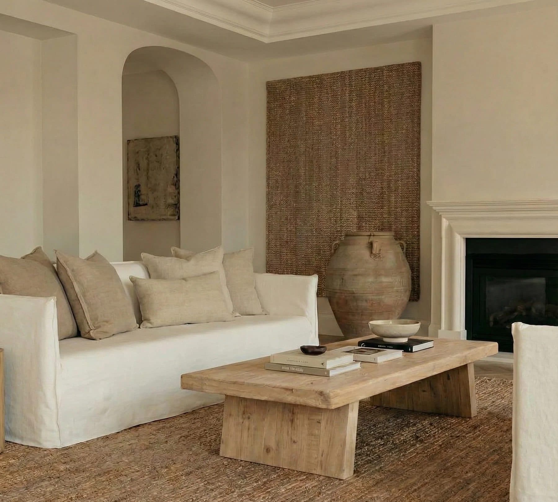 A sunlit living room with a cream linen sofa, cushions, a rustic clay urn, a wooden coffee table with books and a fireplace.
