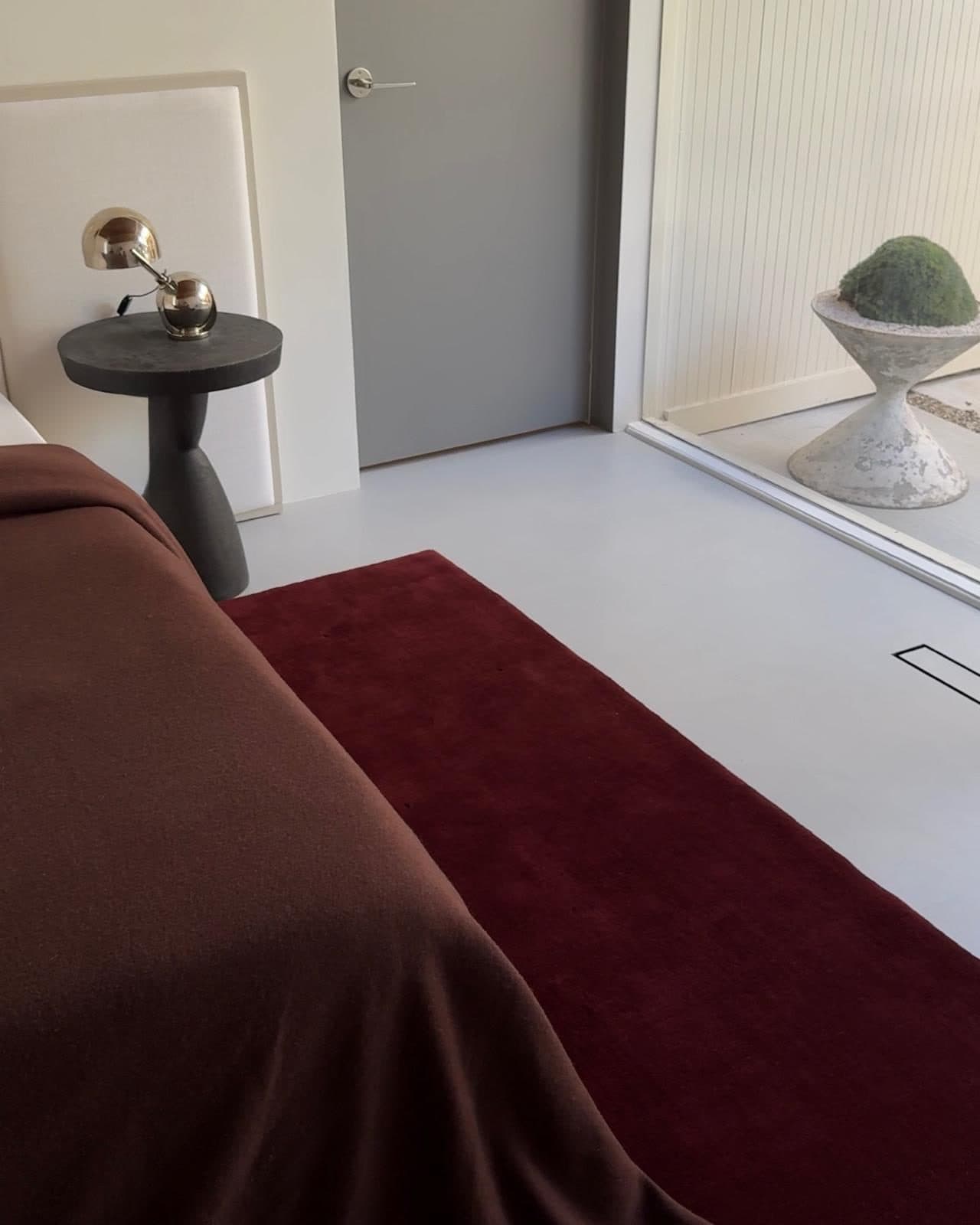 A room with a deep red rug, a brown velvet chair, a mirror and a black side table.