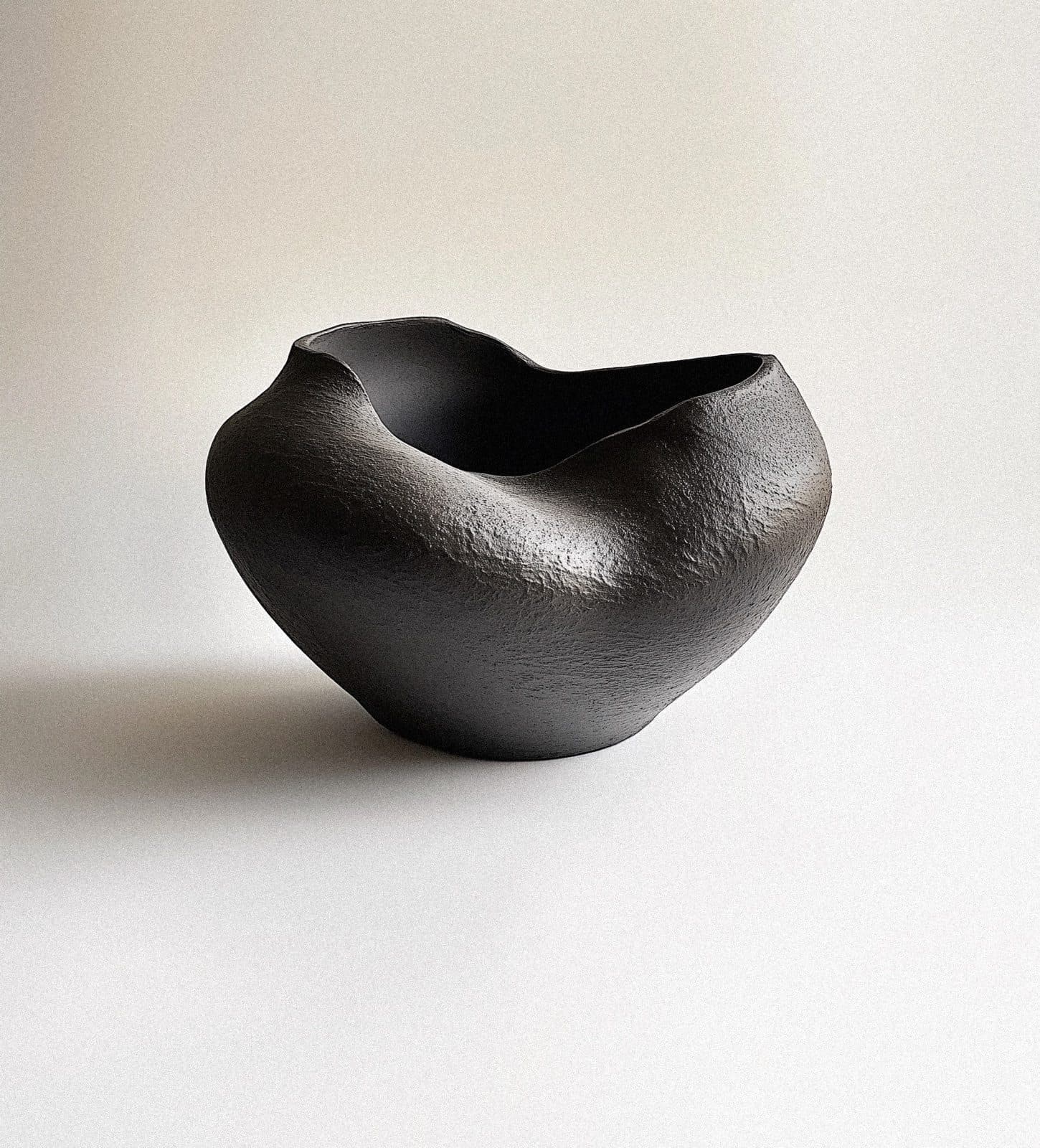 A dark, rough-textured ceramic bowl with a wavy rim on a pale ground, in soft light.