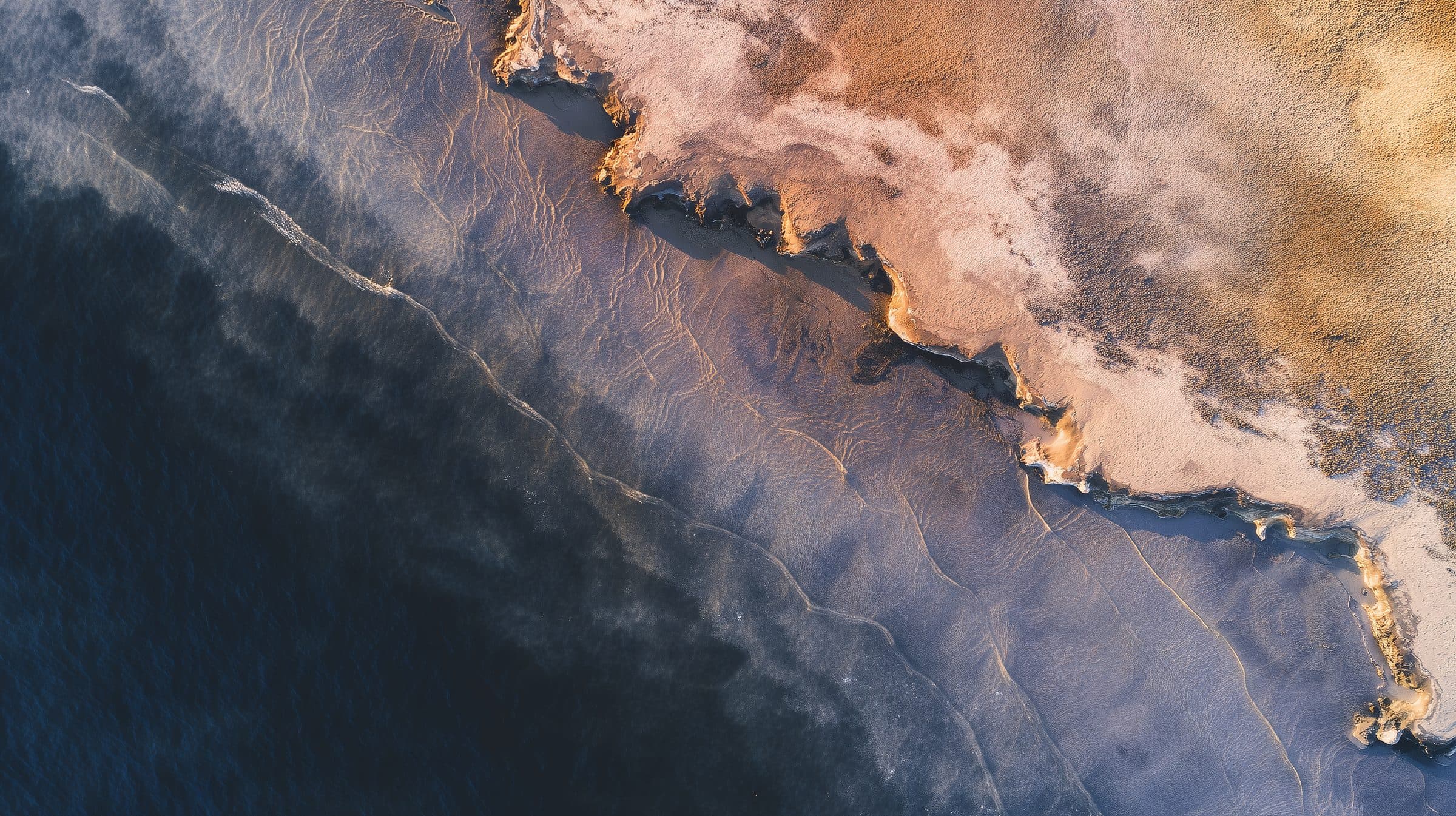 An aerial view of a pale, cracked coastline meeting dark blue water, with faint tracks in the surf.