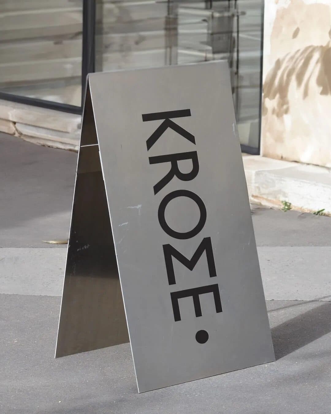 A steel A-frame sign with the Krome wordmark in black, standing on a pavement beside a glass shopfront.