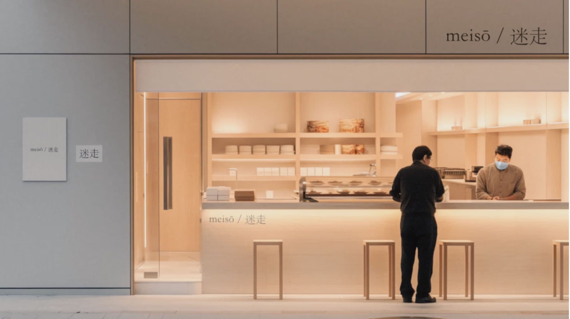 A warmly lit counter at the back of a pale room, with shelves of bread behind it and two people standing at it.