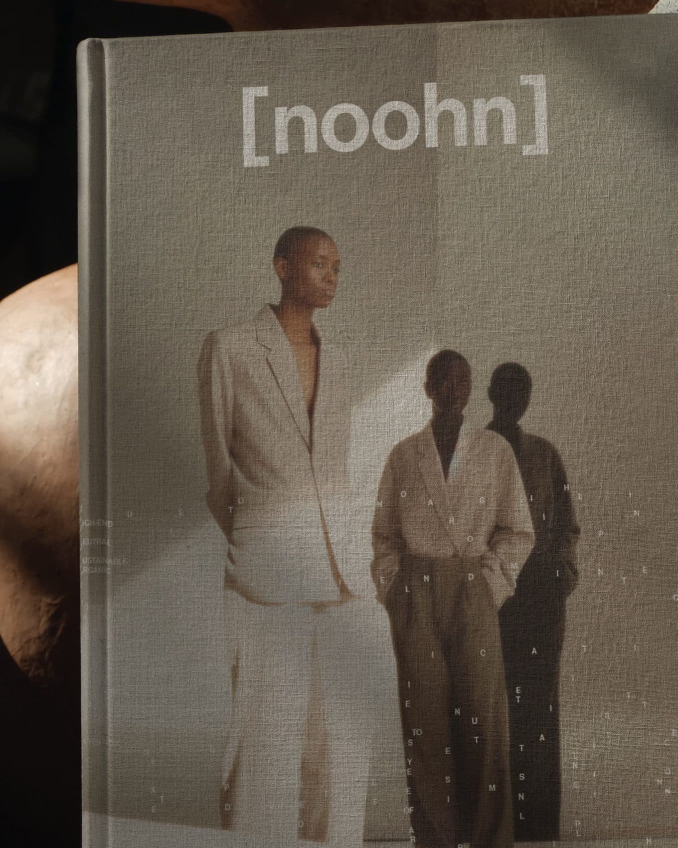 Two people in neutral tailoring beside a shadow, on the cover of a grey linen book marked noohn.