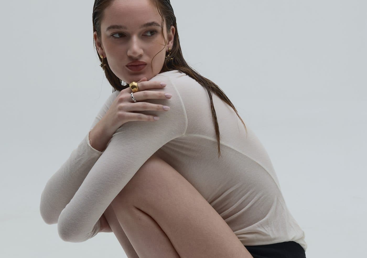 A model in a cream top crouches and hugs her knees, looking over her shoulder, wearing gold rings and drop earrings.