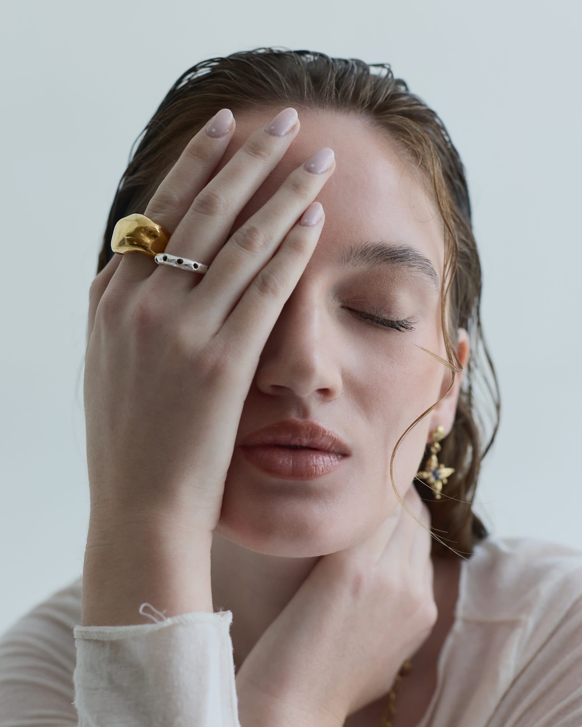 A model with wet-look hair covers one eye with her hand, wearing two chunky rings and a star-shaped earring, against a pale blue-grey wall.