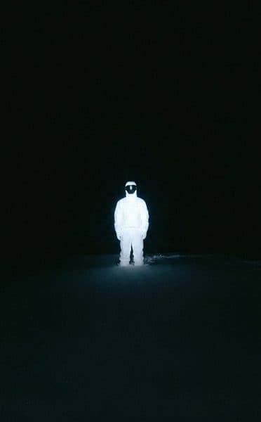 A lone figure in white light standing in a black field.