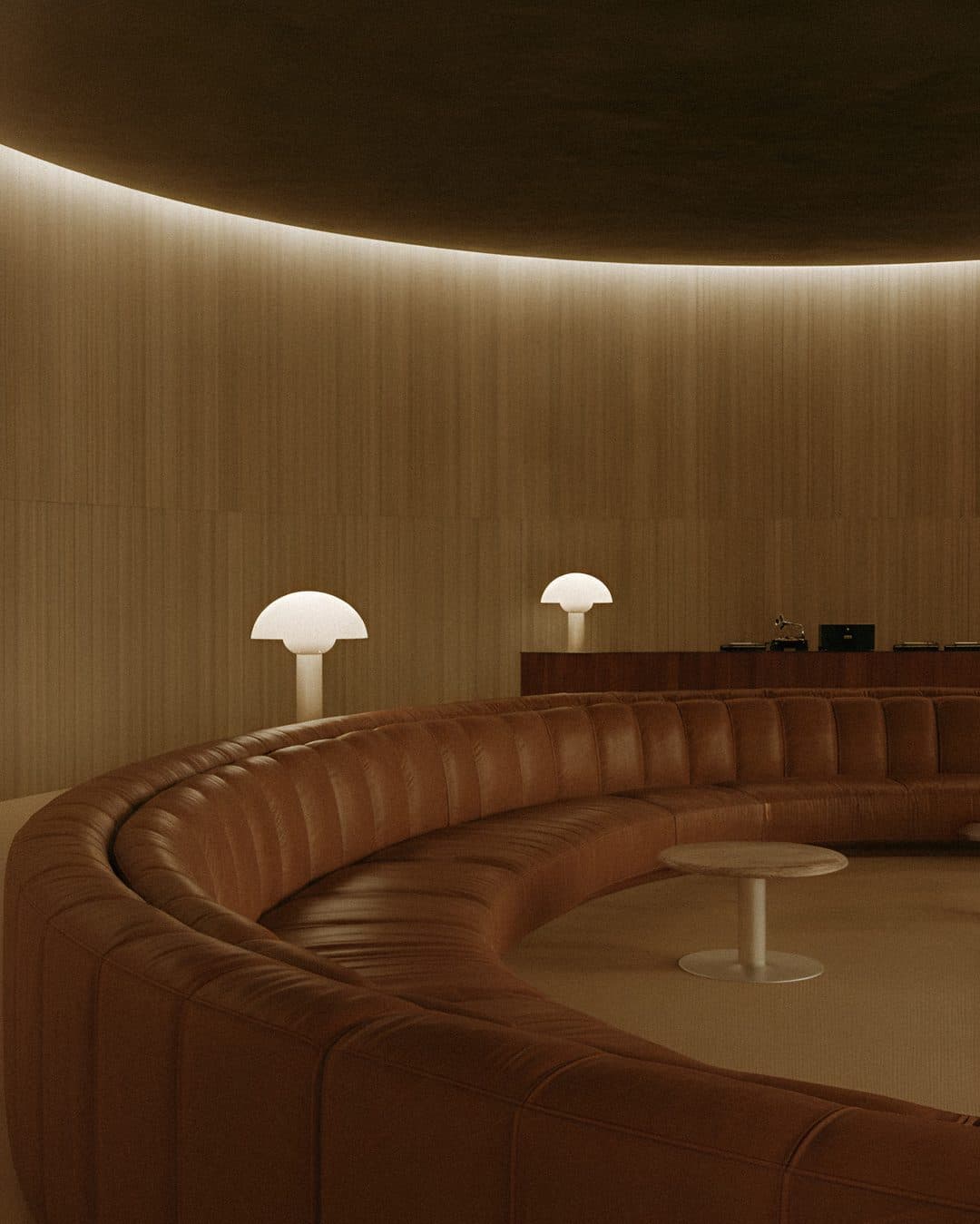 A curved tan leather banquette sweeping around a round, timber-panelled room lit by a soft ceiling glow and two mushroom lamps.
