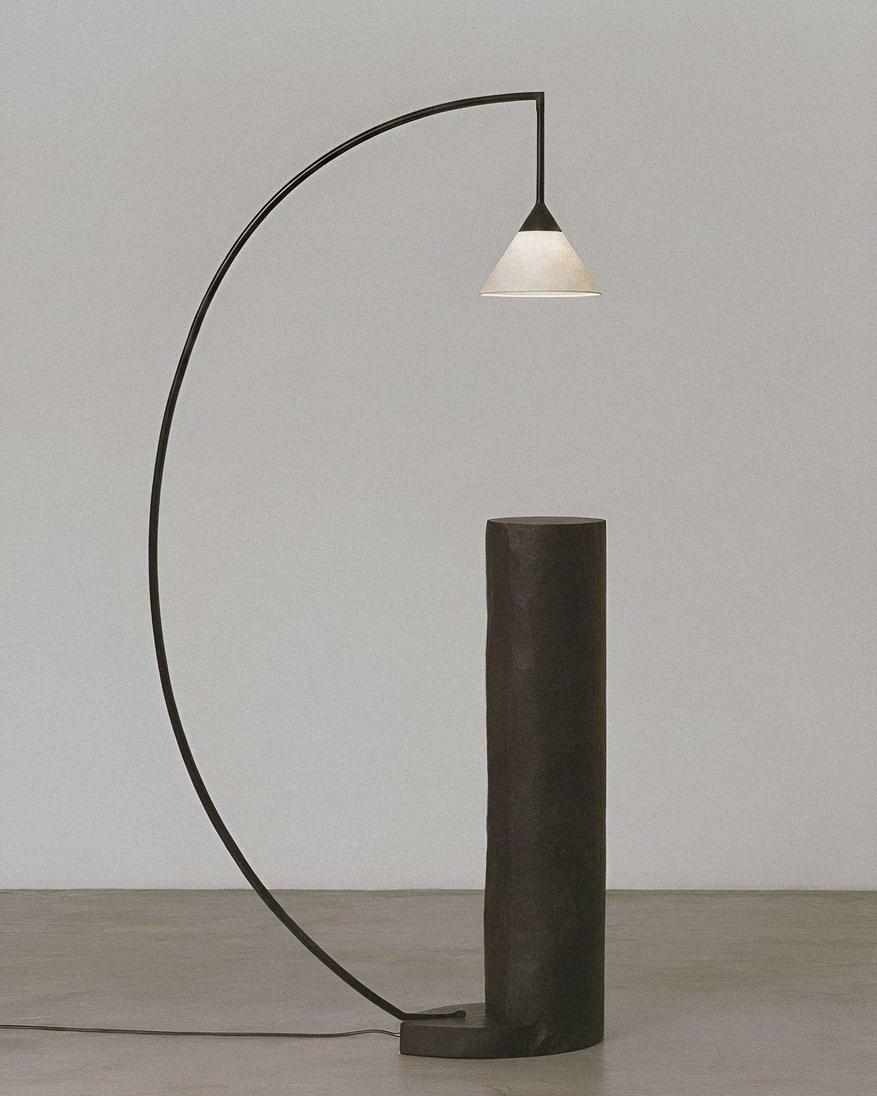 A slim black arc floor lamp with a small pale shade curves over a tall dark cylinder, in a bare grey room.