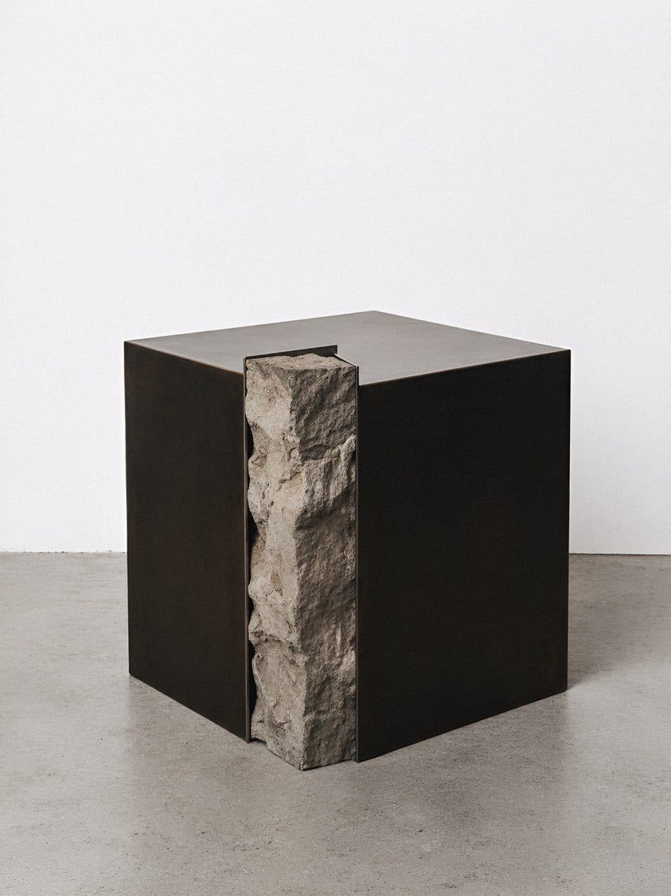 A black steel cube side table with a strip of raw stone set into one face, on a pale concrete floor.