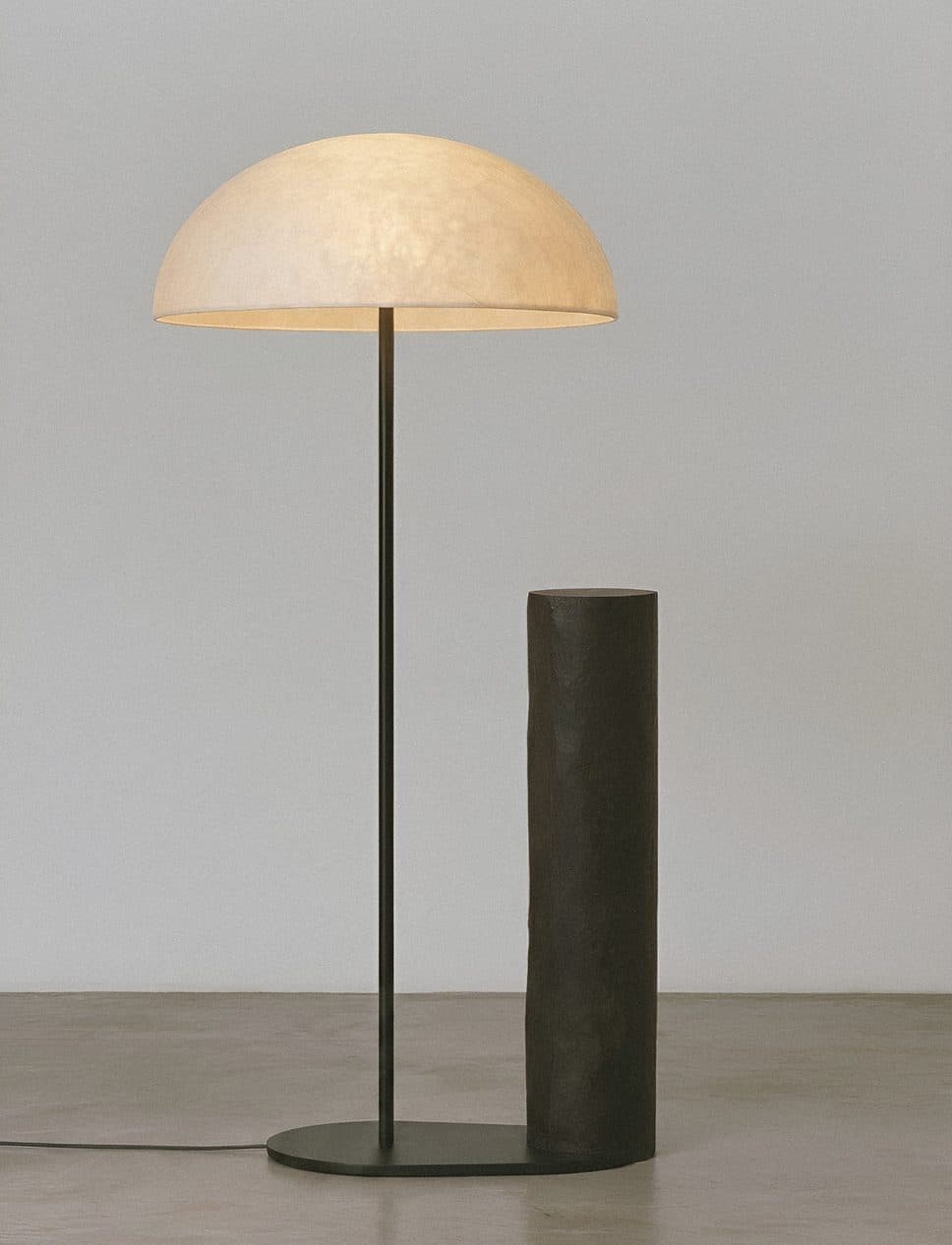 A tall floor lamp with a pale dome shade beside a dark cylindrical base, in a bare grey room.