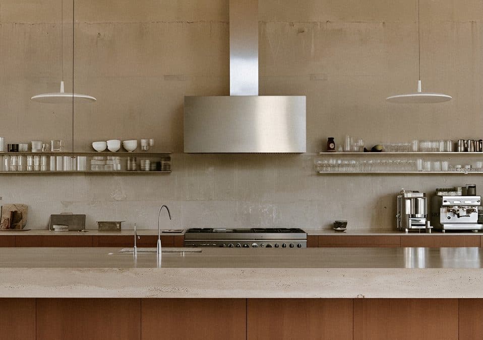 A concrete-walled kitchen with a steel range hood, open shelves of glasses and bowls, and an espresso machine behind a timber counter.