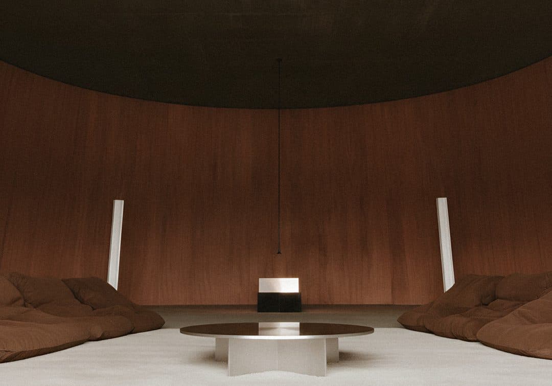 A round timber-panelled room under a dark ceiling, with floor cushions and a low steel table in the middle.