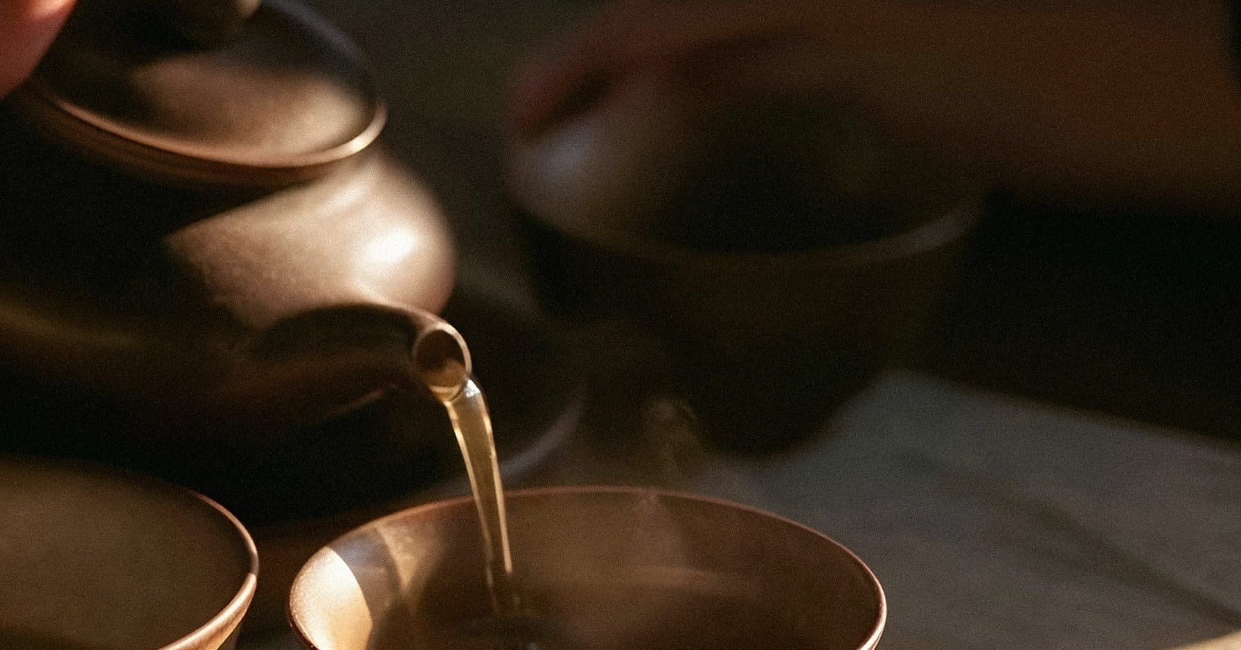 A hand pours tea from a clay teapot into a brown bowl on a sunlit linen-covered table.