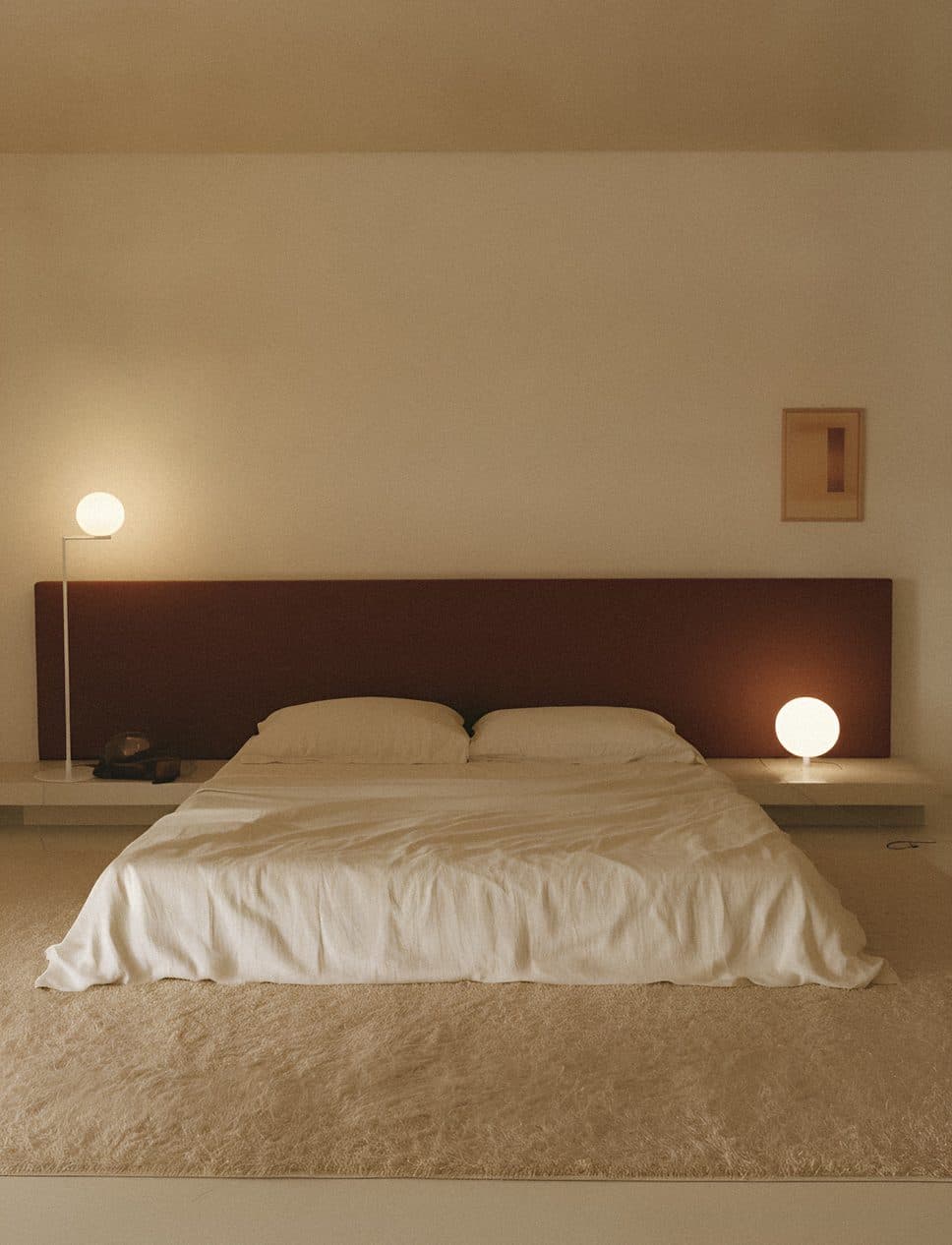 A low bed with white linen and a dark brown headboard between two round lamps in a cream room.