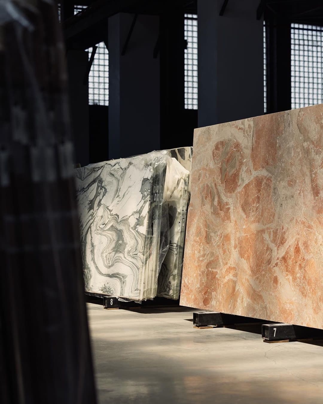 A white marble slab and a stack of stone tiles standing in a warm-lit stone yard, graded dark.