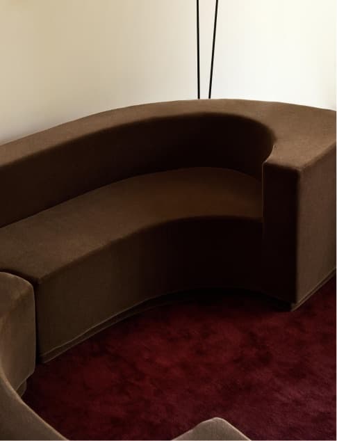 A curved brown velvet sofa in a pale room with a dark red carpet.