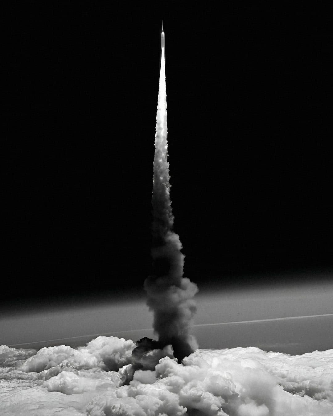 A single rocket rising on a column of white smoke above a bank of cloud, in black and white against a black sky.