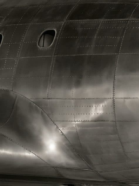 Riveted, dark polished aluminium panels of an aircraft fuselage with a small porthole.