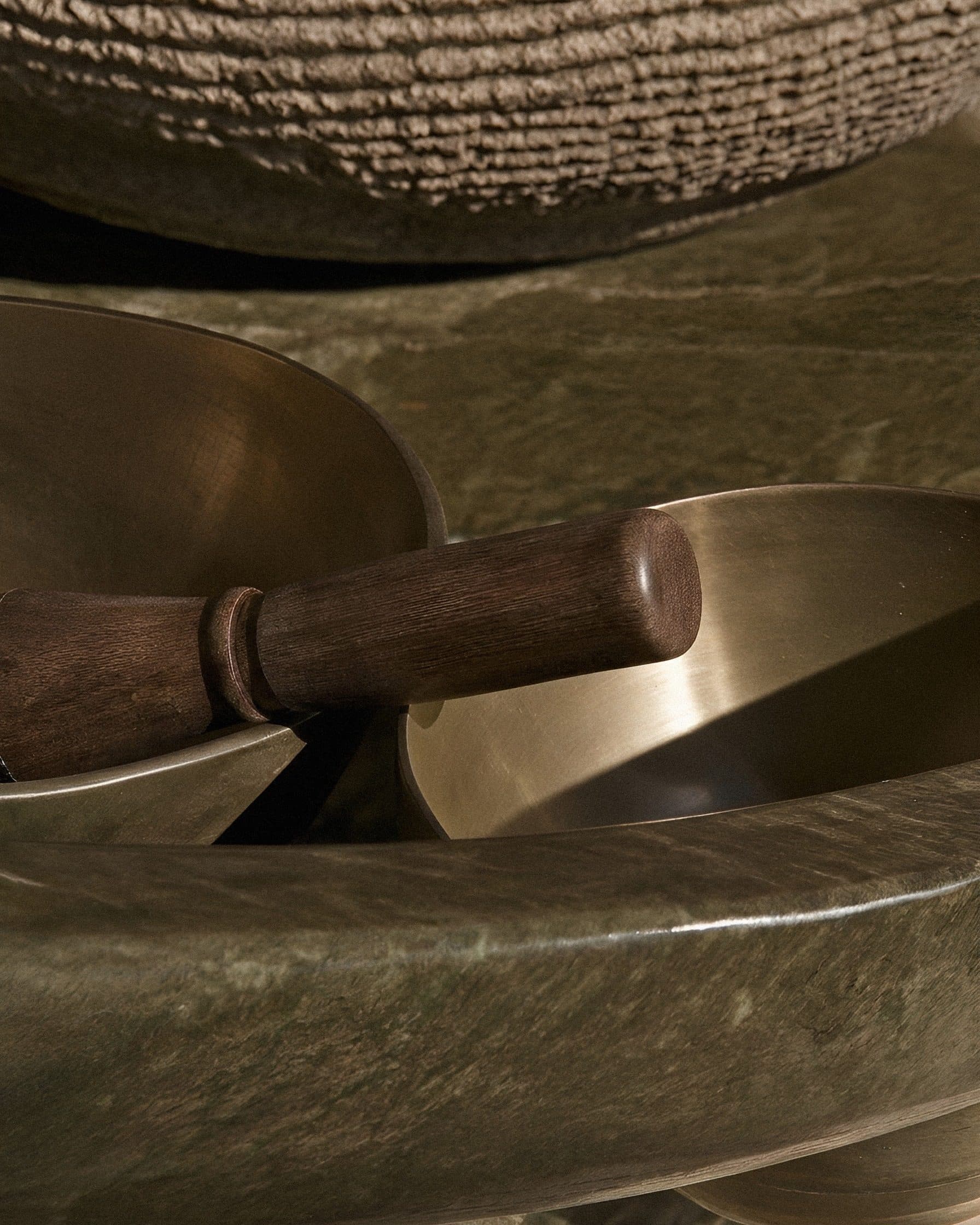A wooden mallet resting across two brass singing bowls on a green stone ledge, in low warm light.
