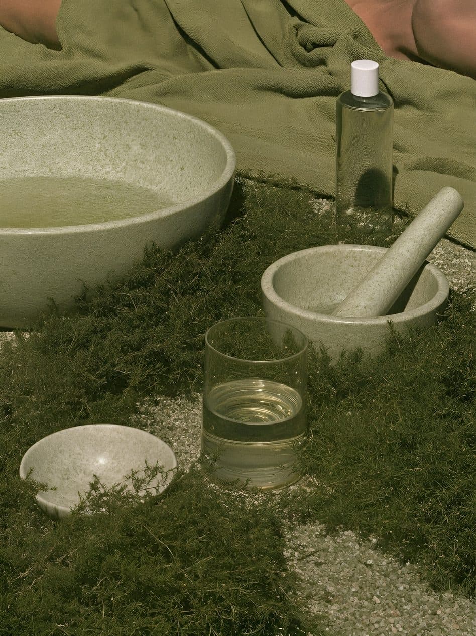 A glass bottle, a glass of water, a stone bowl and a pestle laid out on green moss beside a pale basin.
